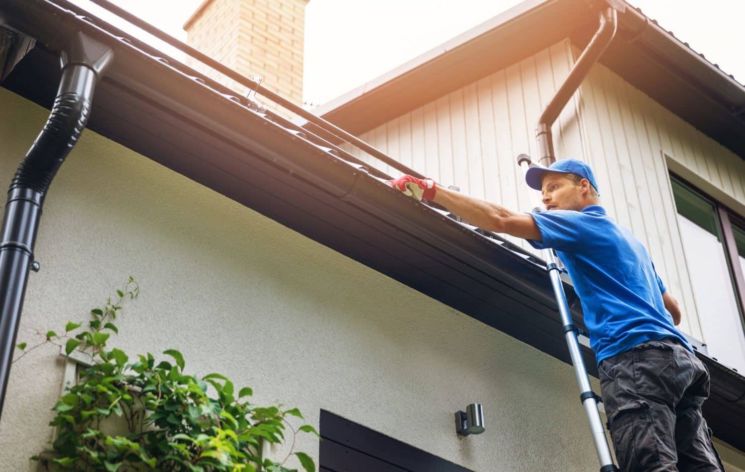 Man on ladder cleaning gutters in Dublin OH. Homeowner wearing blue shirt and cap clears debris from dark gutters on a light-colored house. Professional gutter cleaning in Dublin OH.