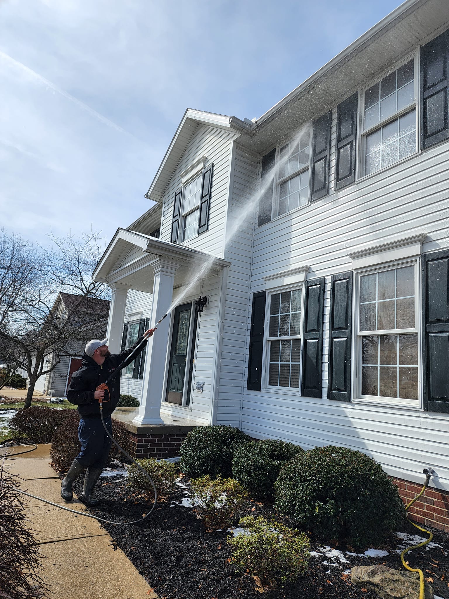 A man power washing a white house with black shutters. This house washing project in Dublin, OH, removes dirt and grime, improving curb appeal.